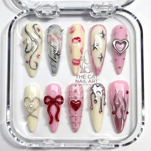 以下が含まれることがあります： 透明なプラスチックケースに入った10本のつけ爪のセット。爪には、シルバーの渦巻き、唇のプリント、星、ハートなど、さまざまなデザインが施されています。ピンク色の爪もあれば、クリーム色の爪もあり、「Angel」の文字が入った爪もあります。