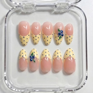 Peut inclure: Un ensemble de dix faux ongles dans un étui en plastique transparent. Les ongles présentent une base rose clair avec un motif à pois jaunes sur les pointes. Certains ongles ont un motif de myrtille.