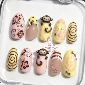 May include: A set of ten artificial nails with various designs. The nails feature a mix of pink, yellow, and brown colors. Some nails have floral patterns, while others have monkey figures or spiral designs. Rhinestones and star-shaped accents are also included.