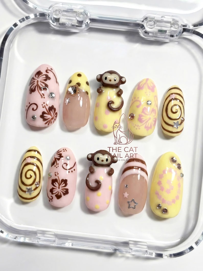 May include: A set of ten artificial nails with various designs. The nails feature a mix of pink, yellow, and brown colors. Some nails have floral patterns, while others have monkey figures or spiral designs. Rhinestones and star-shaped accents are also included.