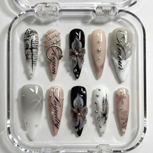 May include: A set of ten artificial nails in a clear plastic case. The nails feature various designs, including floral accents, script, and embellishments. Colors include black, white, pink, and gray. Some nails have the words "Amour" and "Loyalty".