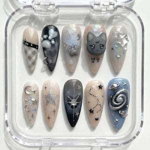 May include: A set of ten almond-shaped false nails in a clear plastic case. The nails feature various designs, including a cat face, stars, and celestial patterns. Colors include beige, black, silver, and blue. Some nails have glitter and small charms.
