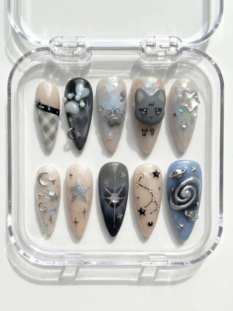 May include: A set of ten almond-shaped false nails in a clear plastic case. The nails feature various designs, including a cat face, stars, and celestial patterns. Colors include beige, black, silver, and blue. Some nails have glitter and small charms.