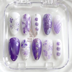 May include: A set of ten press-on nails in a clear plastic case. The nails feature various designs in shades of purple and white, including floral patterns, marble effects, and glitter accents. The text "THE CAT NAIL PRESS-ONS" is visible.