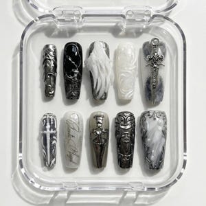 Pode incluir: Um conjunto de dez unhas postiças numa caixa de plástico transparente. As unhas apresentam vários designs, incluindo enfeites prateados ornamentados, padrões em espiral e um motivo de espada. As cores incluem preto, branco, cinza e prata.