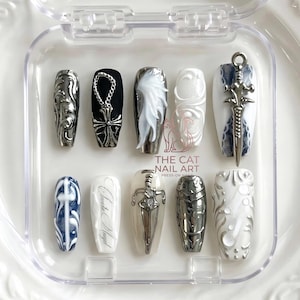 Op de afbeelding: Een set kunstnagels in een doorzichtige plastic doos. De nagels hebben verschillende ontwerpen, waaronder zilveren versieringen, een zwaard en een kruis. Sommige nagels zijn zwart, wit en zilver. De tekst "THE CAT NAIL ART" is zichtbaar.