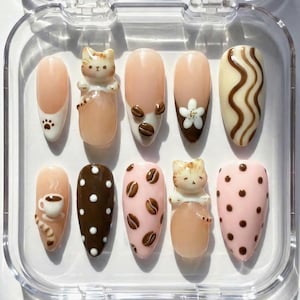 May include: A set of ten artificial nails in a clear plastic case. The nails feature various designs, including a paw print, coffee beans, a flower, a cat figurine, and polka dots. Colors include nude, brown, and white.