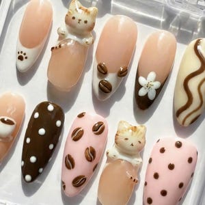 May include: A set of ten artificial nails with various designs. Some nails feature coffee bean and paw print accents, while others showcase a cat figurine, white polka dots, and a flower. The nails are in a clear plastic container.