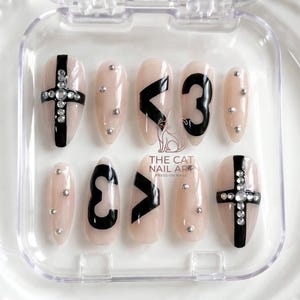 May include: A set of press-on nails in a clear case. The nails are a light pink color with various designs, including black crosses with rhinestones, black chevrons, the number 3, and silver studs. The text "THE CAT NAIL ART" is visible.
