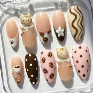 May include: A set of ten artificial nails with various designs. The nails feature a light pink base with designs including coffee beans, a cat figurine, a coffee cup, and polka dots. Some nails have a white base with brown accents.