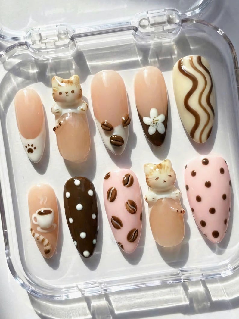 May include: A set of ten artificial nails with various designs. The nails feature a light pink base with designs including coffee beans, a cat figurine, a coffee cup, and polka dots. Some nails have a white base with brown accents.
