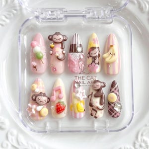 May include: A set of ten almond-shaped press-on nails in a clear plastic case, featuring pink, yellow, and brown designs with 3D monkey figures, bananas, strawberries, musical notes, and piano keys. The text THE CAT NAIL ART is printed in the centre.