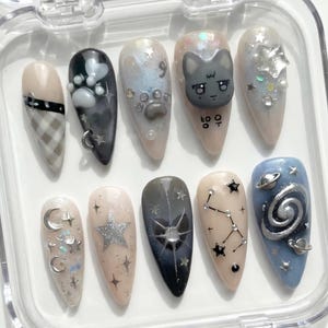 May include: A set of ten artificial nails in a clear plastic case. The nails feature various designs, including a plaid pattern, paw prints, a cat face, stars, moons, and constellations. Colors include beige, gray, black, silver, and blue.