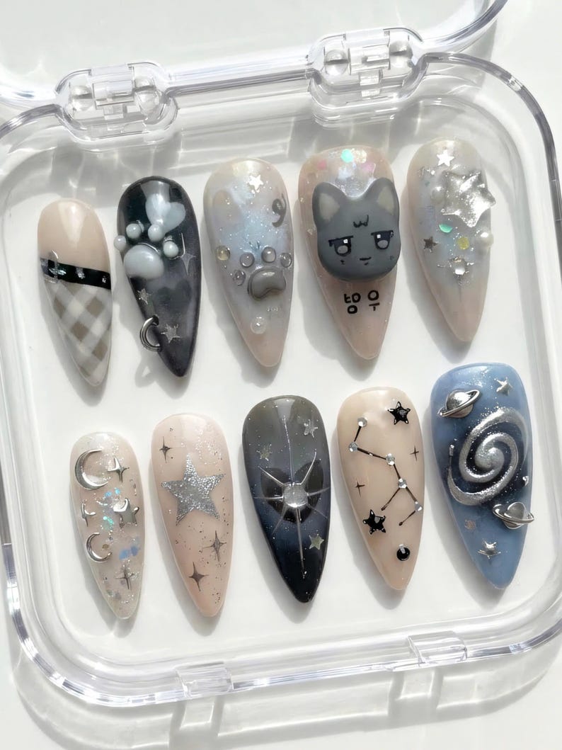 May include: A set of ten artificial nails in a clear plastic case. The nails feature various designs, including a plaid pattern, paw prints, a cat face, stars, moons, and constellations. Colors include beige, gray, black, silver, and blue.