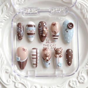 Könnte beinhalten: Ein Set aus zehn Press-on-Nägeln in einer durchsichtigen Kunststoffbox mit der Aufschrift THE CAT NAIL ART. Die Nägel sind in Braun, Beige und Hellblau gehalten und mit weißen Punkten, Noten, Klaviertasten, Schleifen, Knöpfen und 3D-Bären verziert.