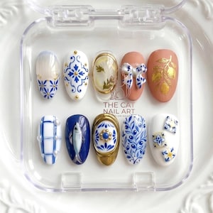 May include: A set of ten artificial nails with various designs. The nails feature patterns such as blue and white floral, a fish, and a bow. Some nails have gold accents and are displayed in a clear plastic case.