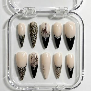 Puede incluir: Un juego de diez uñas artificiales en una caja de plástico transparente. Las uñas presentan una base beige claro con puntas negras y varios diseños, incluyendo piel de serpiente, cruz y patrones arquitectónicos. Algunas uñas tienen pedrería.