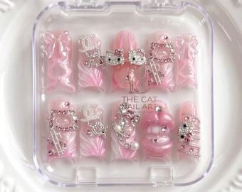 Puede incluir: Un juego de diez uñas postizas en un estuche de plástico transparente, con una paleta de colores rosa claro y blanco. Las uñas están decoradas con lazos en 3D, logotipos plateados, caras de gato con pedrería, perlas y labios rosas brillantes.