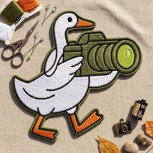 May include: An embroidered patch featuring a white goose with orange feet and beak holding a large olive green camera with a prominent lens. The patch is surrounded by sewing supplies including scissors, thread spools, pins, a film canister, and a small metal camera charm.