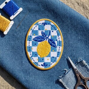 May include: An oval embroidered patch featuring a yellow lemon with blue leaves centered on a blue and white checkered background, bordered by a gold thread edge, placed on blue denim fabric with blue and yellow thread spools and scissors.