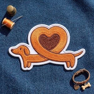 May include: An embroidered patch featuring a brown dachshund dog whose long body curves upward to form a heart shape. The patch has a white border. Nearby are a gold thimble, a spool of orange thread with a needle, and a small brown leather collar.