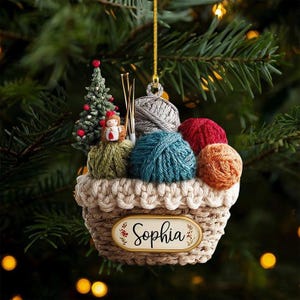 May include: A Christmas ornament shaped like a basket filled with yarn balls in various colors. The basket has a personalized nameplate that reads "Sophia". A small Christmas tree and a figurine are also included in the ornament.