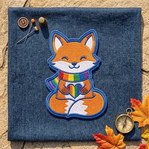 May include: An embroidered patch of an orange fox with white accents, wearing a rainbow-striped scarf and holding a rainbow heart. The patch is placed on dark blue denim fabric alongside a brown button, sewing pins, and a gold-toned compass.
