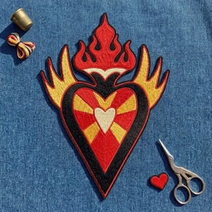 May include: An embroidered patch featuring a central heart with red, yellow, and black stitching, topped with stylized flames and flanked by wings. The patch is accompanied by a metal thimble, a small bundle of colorful thread, a pair of scissors, and a tiny red heart.