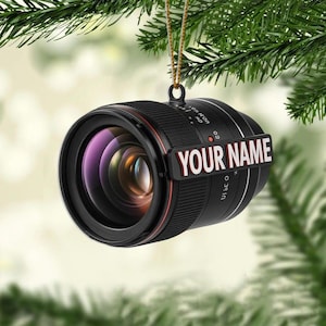 May include: A black camera lens ornament with a red accent ring and the text "YOUR NAME" in white. The ornament is hanging from a gold string against a backdrop of green pine needles.