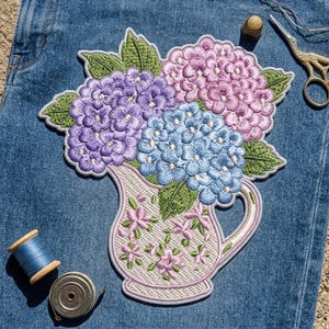 May include: An embroidered patch featuring a bouquet of purple, pink, and blue hydrangea flowers in a white pitcher with a small pink floral pattern. The patch is placed on blue denim fabric alongside a blue thread spool, a metal bobbin, and gold scissors.