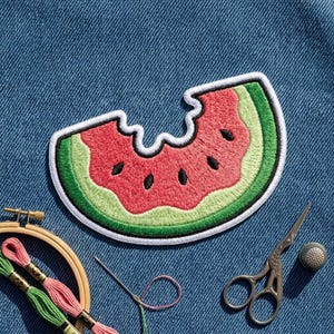 May include: An embroidered patch shaped like a bitten watermelon slice with red fruit, black seeds, and a green rind, outlined in white thread. The patch is placed on blue denim fabric next to embroidery floss, a needle, and a metal thimble.