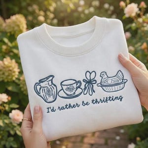 Op de afbeelding: Crèmekleurige sweatshirt met een ronde hals en de geborduurde tekst "I'd rather be thrifting". Het marineblauwe ontwerp bevat een kan, een kopje, een strik en een kip in een mand.