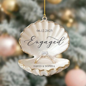 May include: A white seashell-shaped ornament with gold trim, featuring the word "Engaged" in elegant script. It holds a diamond engagement ring. The ornament includes the date 06.12.2025 and the names "DAVID & SOPHIA".
