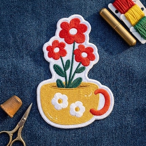 May include: An embroidered patch featuring a yellow teacup with a red handle and two white flowers on the front. Three red flowers with white centers and green stems emerge from the cup. The patch has a white border and is placed on blue denim fabric.
