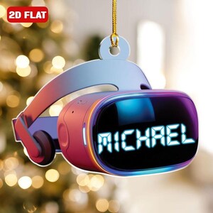 May include: A 2D flat Christmas ornament shaped like a VR headset. The headset is primarily pink and blue with the name "MICHAEL" displayed on the screen. The ornament is suspended by a gold string and has the text "2D FLAT" in the upper left corner.