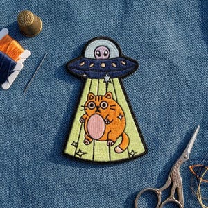 May include: An embroidered patch featuring a dark blue UFO with a light blue dome and a small alien inside. A bright orange cat with a pink belly is being lifted by a light green tractor beam. The patch includes small star details.
