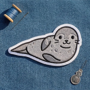 May include: A gray seal embroidered patch with a white border, featuring a spotted pattern, black stitched eyes, and a small nose. The patch is accompanied by a spool of blue thread, a sewing needle, and a metal needle threader tool.