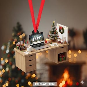 May include: A Christmas-themed ornament shaped like a desk with a laptop displaying the name "BARY". The desk is decorated with a small Christmas tree, gifts, and a mug. The ornament is suspended by a red ribbon.