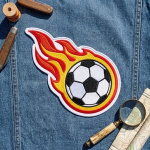 May include: An embroidered patch featuring a black and white soccer ball surrounded by stylized yellow and red flames. The patch has a white border and is positioned on a blue denim fabric background, accompanied by a wooden spool and a magnifying glass.