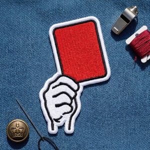May include: An embroidered patch featuring a white hand holding a solid red rectangular card with a thick black border. The patch is displayed on a blue denim fabric background alongside a silver metal whistle, a spool of red thread, and a gold button.