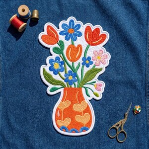 May include: An embroidered patch featuring a bouquet of colorful flowers in an orange vase with heart patterns. The flowers include red tulips, blue daisies, and small pink blossoms with green leaves. A red spool of thread, a thimble, and scissors are nearby.