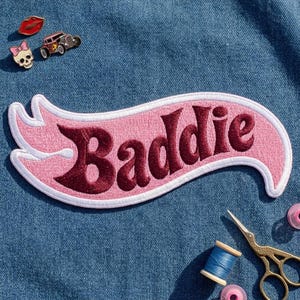 May include: A pink embroidered patch shaped like a banner with the word Baddie in dark red lettering. The patch features a white border and is placed on blue denim fabric alongside small enamel pins, a spool of blue thread, and gold scissors.