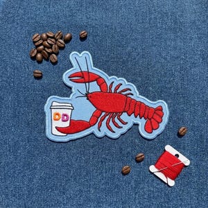May include: An embroidered patch featuring a bright red lobster holding a white coffee cup with the orange and pink letters DD printed on the side. The patch has a light blue border and is surrounded by scattered dark brown roasted coffee beans.