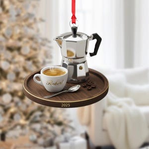 May include: A coffee-themed Christmas ornament featuring a silver espresso maker, a white coffee cup with "Robert" in gold script, and a small spoon on a wooden tray. The tray also has the year "2025" printed on it. A red ribbon is attached for hanging.