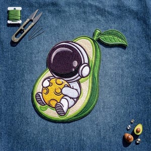 May include: An embroidered patch featuring an astronaut in a white suit holding a yellow moon, positioned inside a green avocado half with a single green leaf. Nearby are metal embroidery scissors, a spool of green thread, and small avocado pieces.