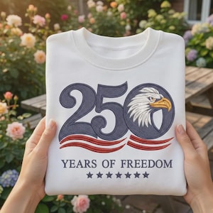 Może przedstawiać: Biała bluza z granatowym wzorem "250" i grafiką głowy orła. Tekst "YEARS OF FREEDOM" i gwiazdy znajdują się poniżej. Bluza ma okrągły dekolt.