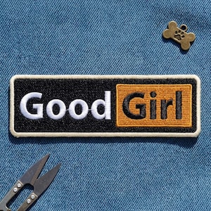 May include: A rectangular embroidered patch with a black background and a cream border. The text 'Good' is in white, and 'Girl' is in black on a mustard yellow rectangular field. A small bronze-colored bone-shaped charm with a paw print is included.