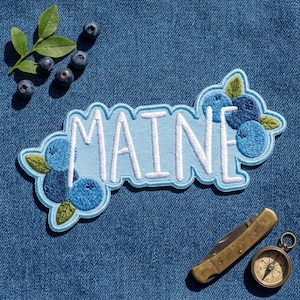 May include: An embroidered patch featuring the word MAINE in white letters on a light blue background, decorated with blue blueberry clusters and green leaves, accompanied by loose blueberries and a brass pocket knife with a compass.
