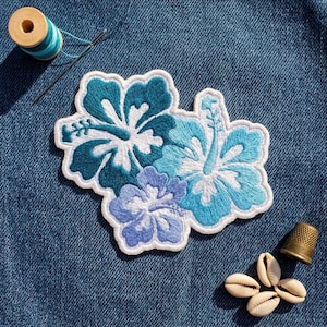 May include: An embroidered patch featuring three hibiscus flowers in shades of dark teal, light blue, and periwinkle with white centers. The patch is placed on blue denim fabric alongside a wooden spool of blue thread, a needle, and four seashells.