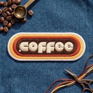 May include: An oval embroidered patch featuring the word coffee in cream-colored lettering centered within concentric bands of brown, orange, and yellow. The patch is surrounded by scattered roasted coffee beans, a metal scoop, and scissors.